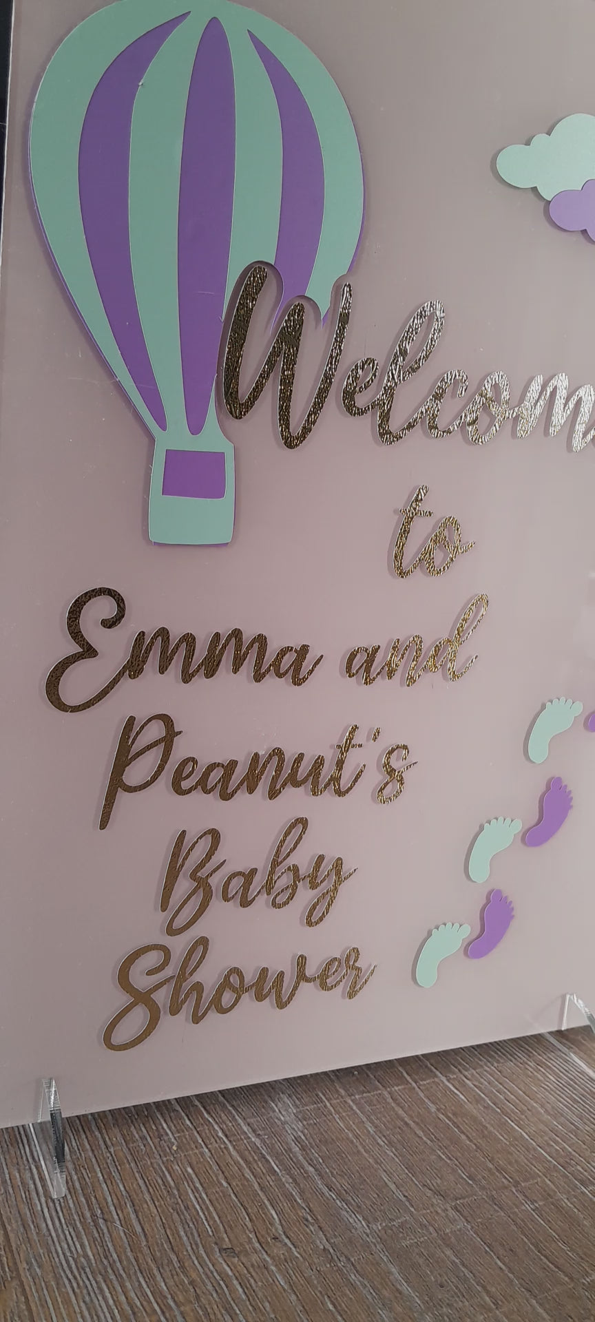 Pink blush baby shower sign with purple and green balloon and clouds. Displays names in gold. 